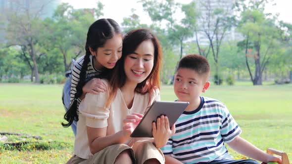 Asian family with two children happy and enjoy sitting using a digital tablet together in park