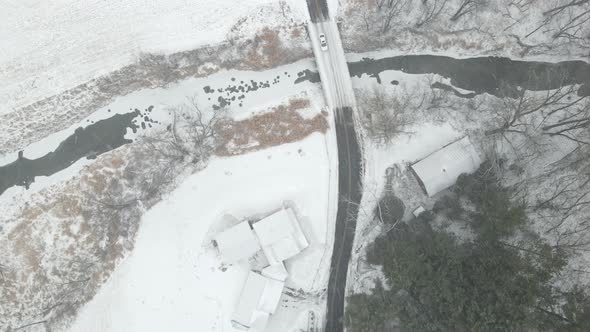 Drone view of lone car on snowy country road in Wisconsin. Bridge over creek running through farm.