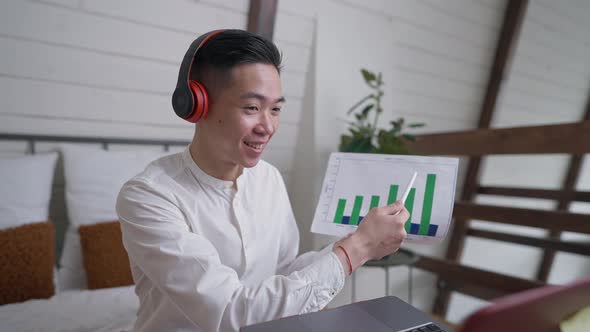 Smiling Chinese Young Man in Headphones Pointing at Graph Talking Using Virtual Conference on Laptop alt