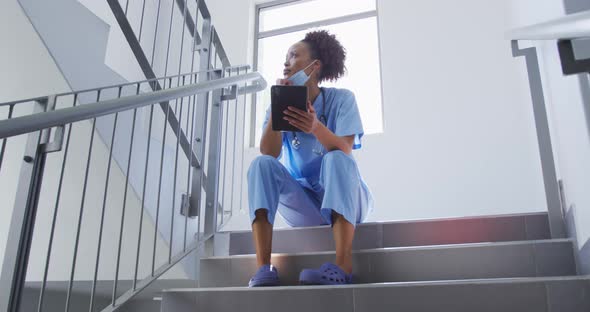 Mixed race female doctor wearing face mask sitting on stairs in hospital using tablet alt