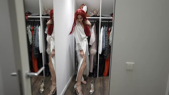 Wide Shot Thoughtful Transgender Woman in White Silk Bathrobe and Red Wig Trying on Luxurious alt