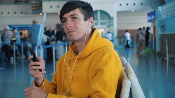 Young Male Hipster in Yellow Hoodie Waiting Airplane in Airport and Using His Smartphone with