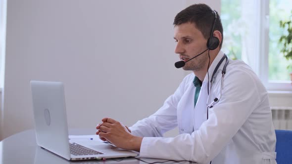 Friendly Male Doctor in White Medical Coat With Headphones Making Conference Call on Laptop alt