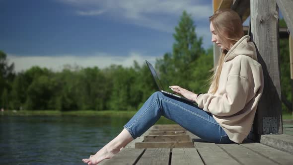 Beautiful Young Girl is Using Laptop and Typing While Sitting on Pier By Lake on Summer Day Spbd alt
