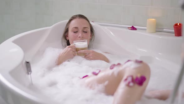 Slim Relaxed Caucasian Woman Lying in Bath Foam in Bathtub Smelling Tasty Cappuccino alt