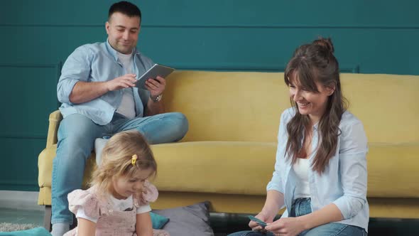 Little Girl Playing with Mother on the Carpet While Her Father Works Using a Tablet alt