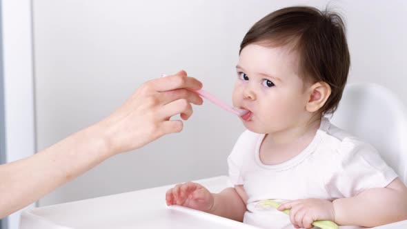 Baby Girl Eating Blend Mashed Food Sitting on High Chair Mother Feeding Child Hand with Spoon for alt