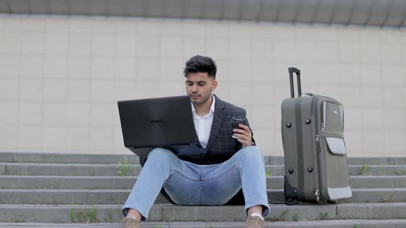 Handsome Arabian Man in Stylish Wear Sitting on Stairs and Working on Wireless Laptop While Waiting alt