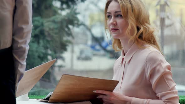 Cheerful Young Women Sitting at Cafe Holding Menu Card Giving Order To Waiter alt
