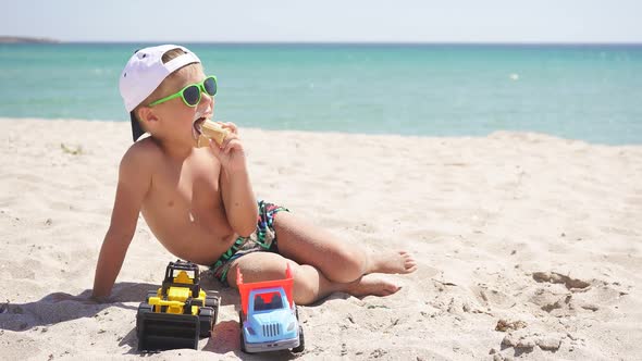 Tanned Boy Wearing a Baseball Cap Eats Ice Cream While Sitting on a Sandy Beach, Against a Beautiful alt
