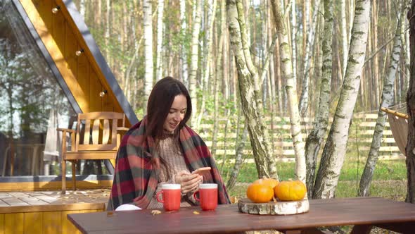 Happy Young Woman with Phone and Drinking Coffee Sitting at the Wooden Table Outdoors