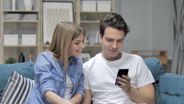 Young Couple Using Smartphone While Relaxing on Couch Messaging alt