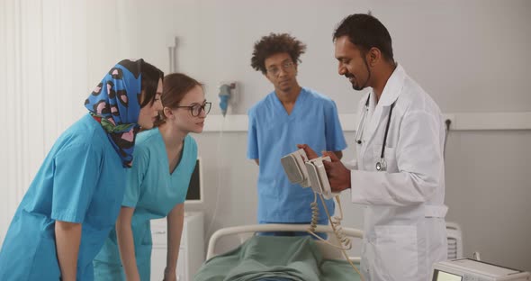Mature Indian Doctor Showing Young Interns How to Use Defibrillator in Hospital Ward alt
