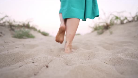 Legs of Woman Wearing Blue Skirt Walking Barefoot on Sandy Beach or Desert. Slow Motion. Girl Walks alt