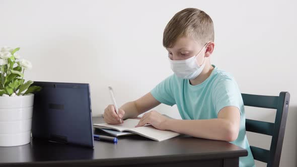 A Young Boy in a Face Mask Does Homework for School with the Help of a Tablet at a Table at Home alt