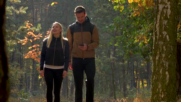 A Hiking Couple Walks Through a Forest on a Sunny Day and Looks at a Smartphone alt