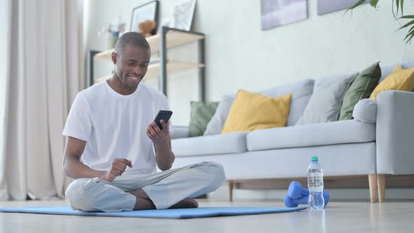 Young African Man Using Smartphone on Yoga Mat at Home alt