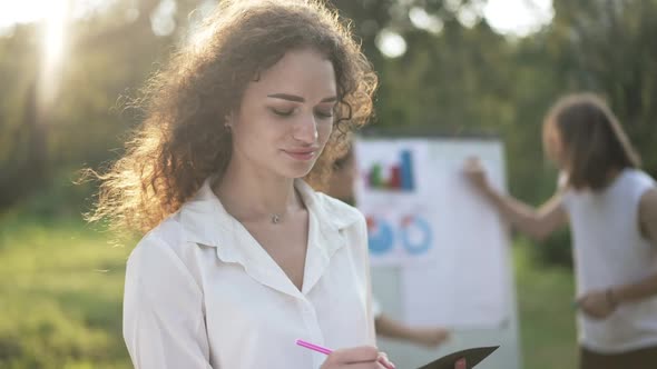 Beautiful Confident Woman Writing with Pen Smiling Looking at Camera As Blurred Colleagues alt