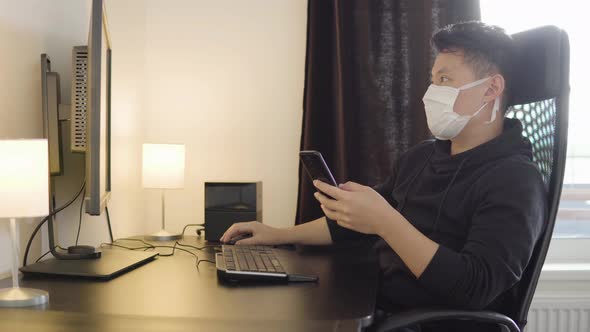 A Young Asian Man in a Face Mask Works on a Desktop Computer and a Smartphone at Home alt
