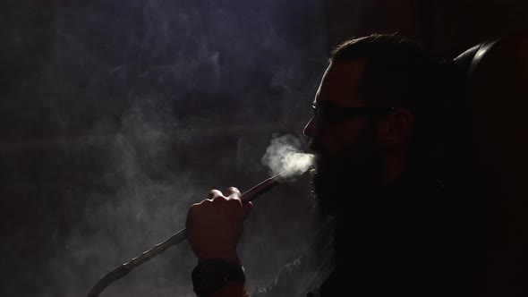 Young Bearded Man in Glasses Smokes a Hookah and Blow Out Smoke Closeup on Black Background in Slow