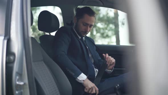 Side View Handsome Caucasian Young Man Fastening Seat Belt Sitting on Driver's Seat in Car alt