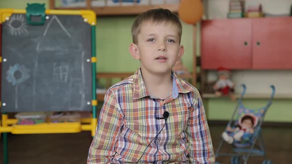 A Boy in a Plaid Shirt Sits on a Chair in Front of the Camera with a Microphone on His Clothes and