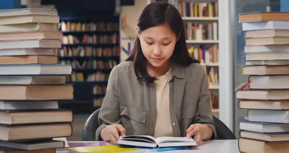 Asian School Student Girl Reading Book Sitting at Desk at Library alt