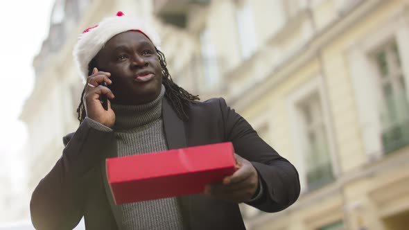 Black Man with Christmas Hat and Red Gift Box Having a Phone Call on the City Street alt