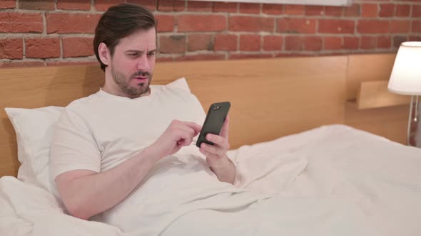 Casual Young Man Reacting to Loss on Smartphone in Bed alt