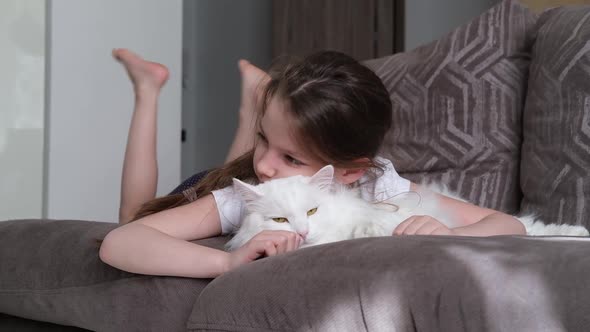 A Little Happy White Girl Lies on the Sofa in the Living Room Hugs a White Fluffy Cat and Smiles