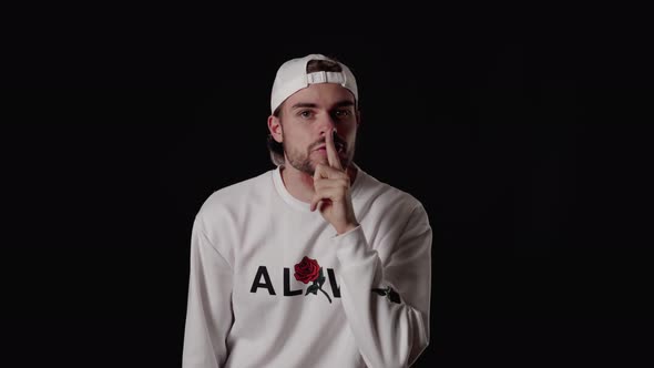 Trendy 20s Man shushing into the camera, shh gesture, wide, black background alt