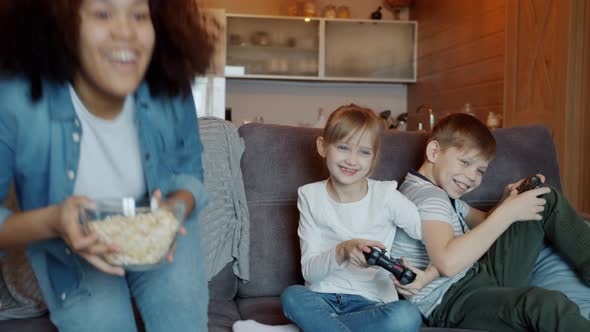 Brother and Sister Playing Video Game When Afro-American Nanny Coming with Popcorn alt
