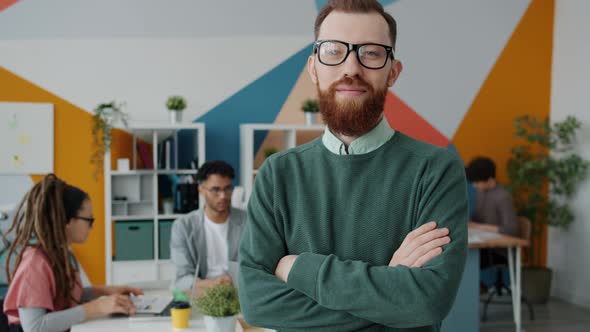 Portrait of Young Man in Glasses Looking at Camera in Office with Colleagues in Background alt