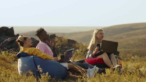 Confident Freelance Team Works Together Using Laptop Sitting Comfortably in Bag Chairs alt