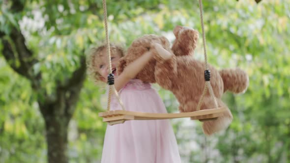 Spring and happy summer time. Joyful smiling little girl playing with teddy bear swinging on the swi