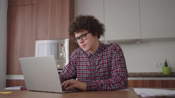 A Curly Man with a Serious Look Works at a Laptop Sitting in a Modern Kitchen alt