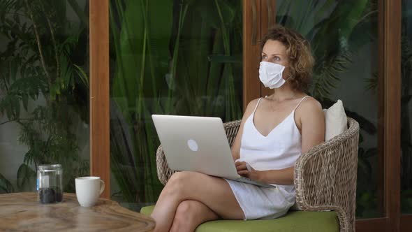 Caucasian Woman Wearing Protective Face Mask Works on a Laptop on a Tropical Terrace alt