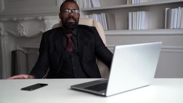 a Bearded AfricanAmerican Man in Glasses and a Suit with a Tie is Sitting at a Table with a Phone alt