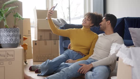 Couple In Lounge Taking Selfie On Mobile Phone With Unpacked Boxes In New Home On Moving Day