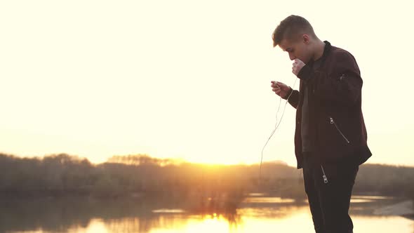 Young Handsome Man Wearing Headphones Listens to Music on Beach During Sunset