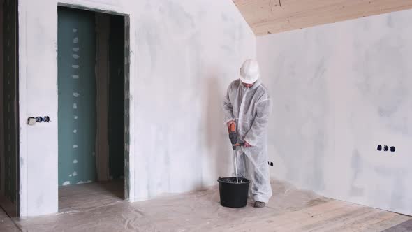 A Man in a Protective Suit and a Hard Hat Mixes Cement for Repairs in Houses