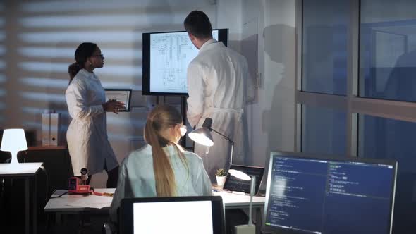 Multiracial Specialists in Electronics Talking Something and Showing on a Big TV Screen in Lab alt