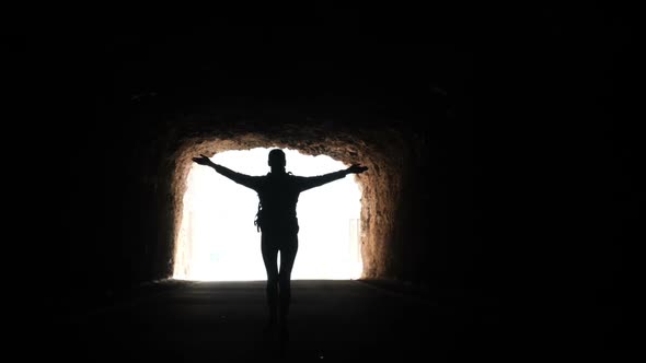 Camera Follows a Woman Silhouette Walking Through Dark Tunnel Towards the Light Raising Arms in Air alt