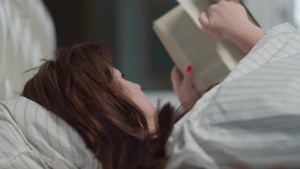 Pretty Brunette Woman Lies in Bed and Reads a Book in the Evening