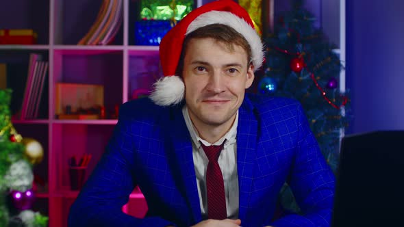 Young Man in Santa Hat Looking at Camera and Sitting in Office alt