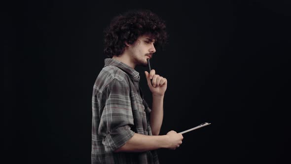 The Attractive Boy with Curly Hair in Profile Thinks Writes Notes with the Pen on the Sheet alt