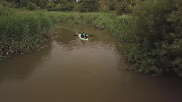 Man kayaking on meandering river with reeds bush’s and trees on either bank. Drone follows behind th alt