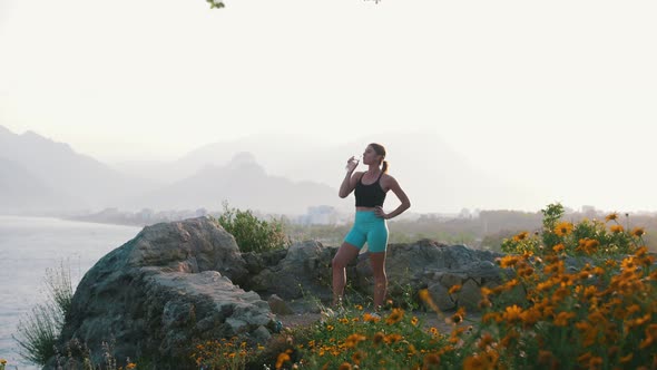 A Woman in Sportswear Drinks Water on a Hill From the Sea alt