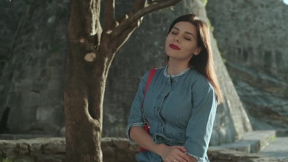 Young smiling brunette woman with red lipstick in denim overalls  sits on sunlights alt