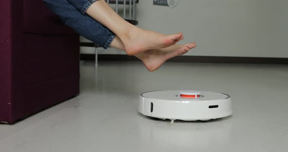 robot vacuum cleaner driving around barefoot woman at home. Female lift feet up when a smart vacuum 